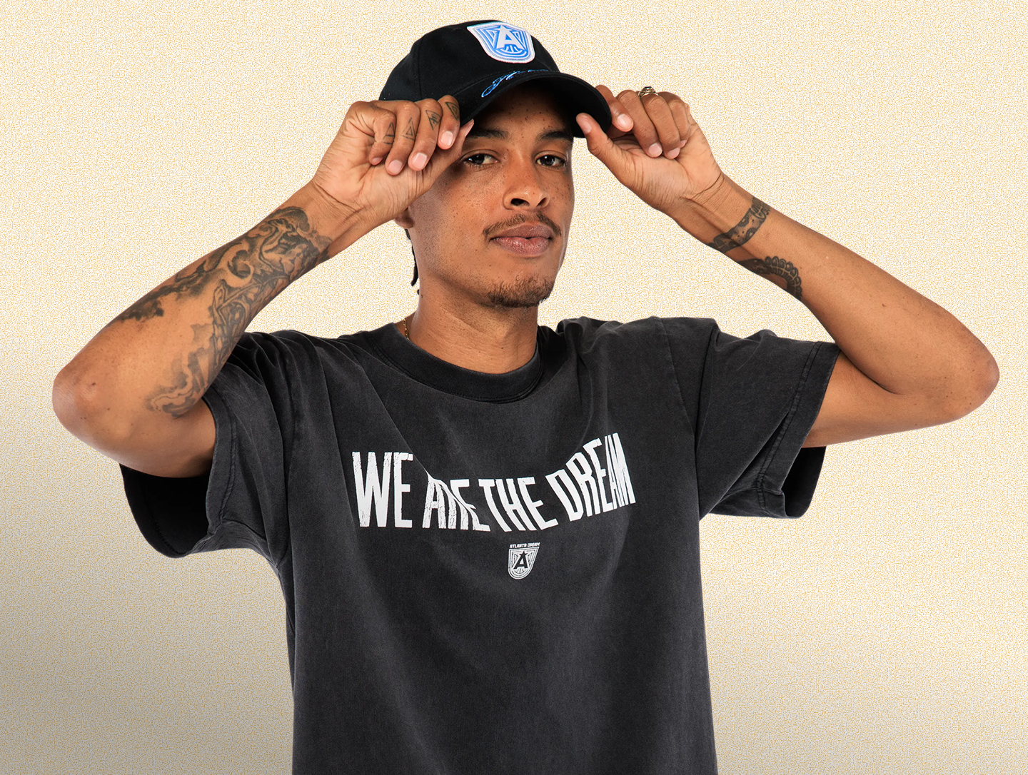 Person wearing a black t-shirt with 'WE ARE THE DREAM' text and a cap on a beige background