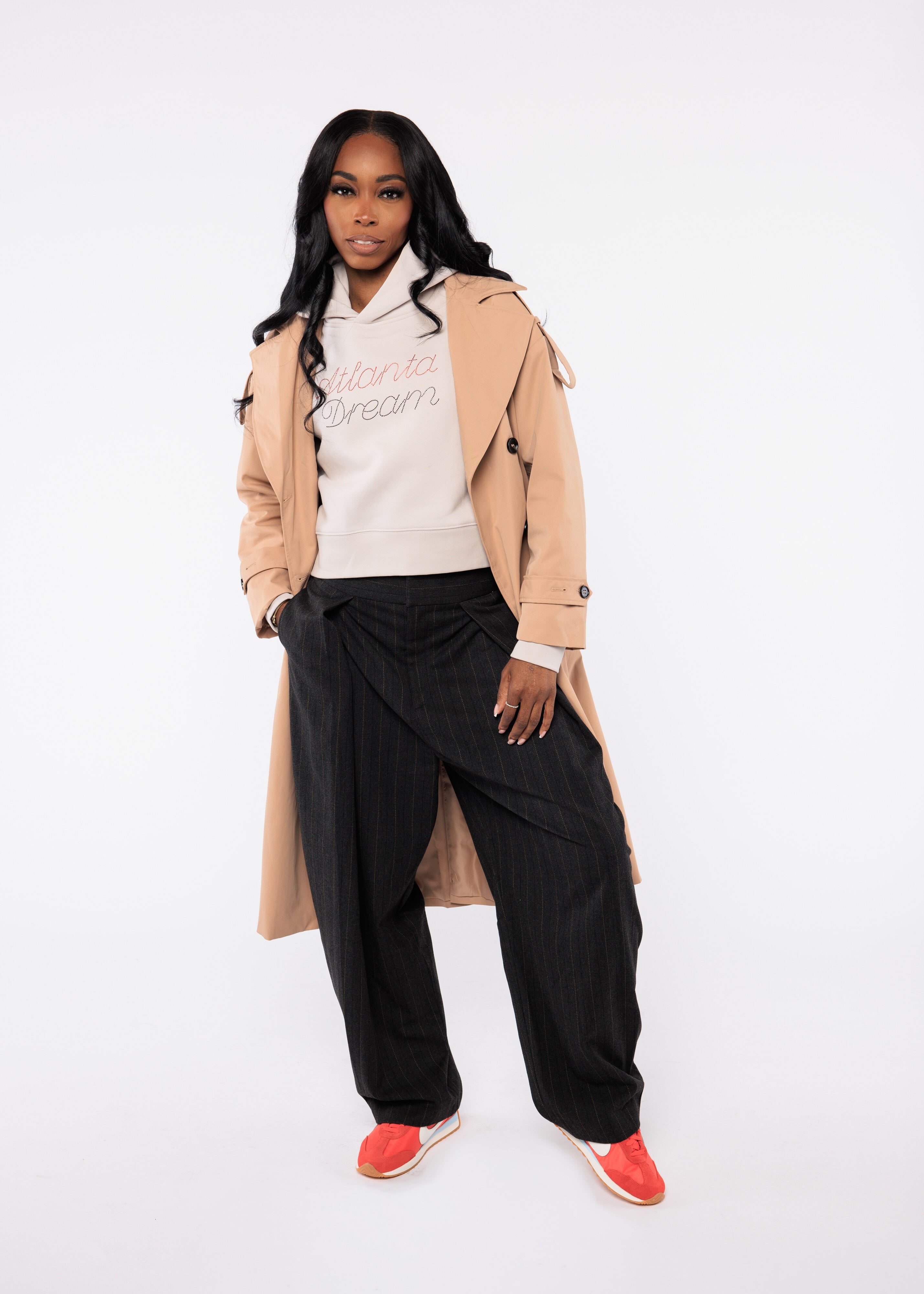 Woman wearing a beige trench coat over a white hoodie with text, black pants, and red shoes