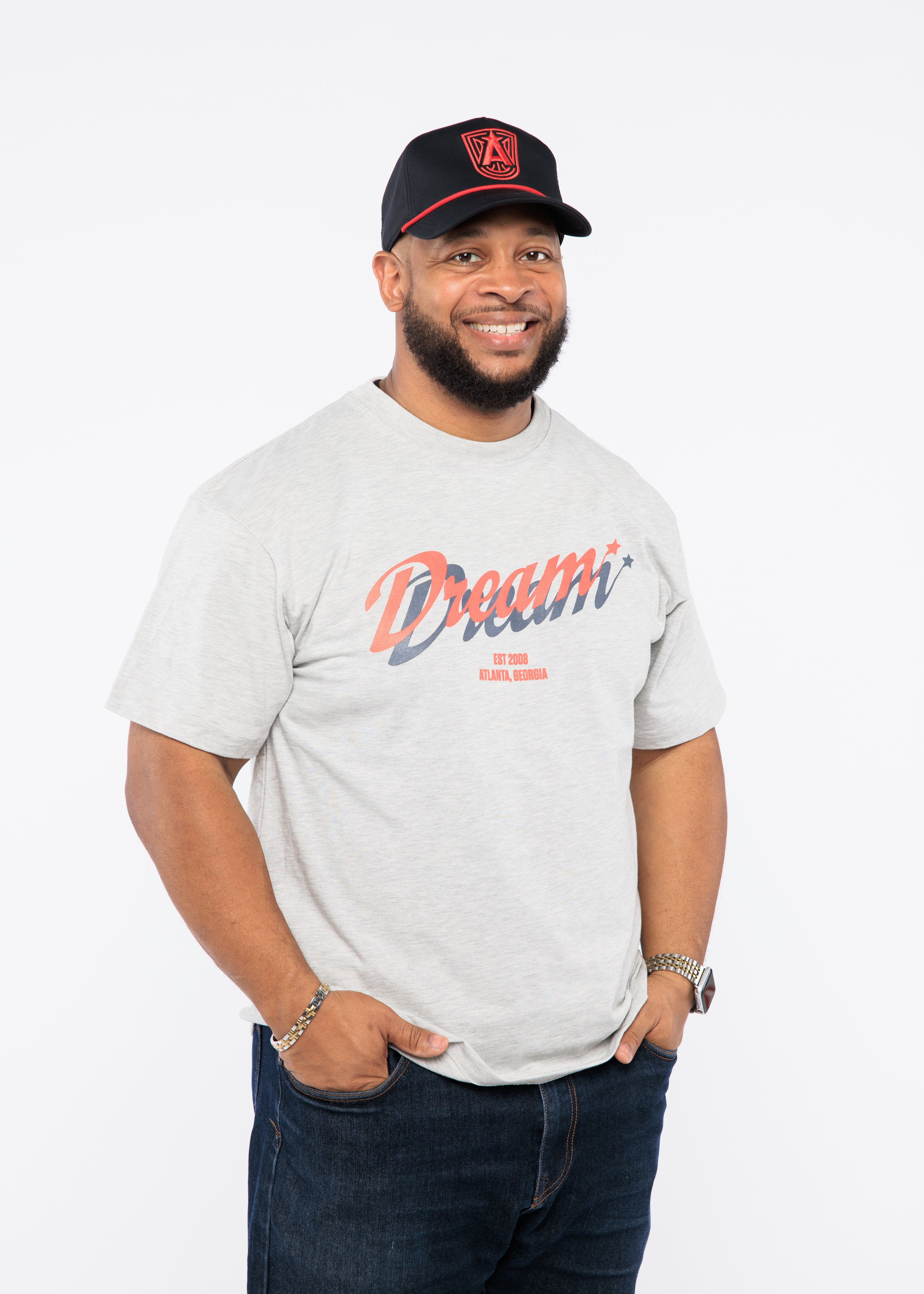 Man wearing a gray t-shirt with 'Dream' printed on it, black cap, and dark jeans