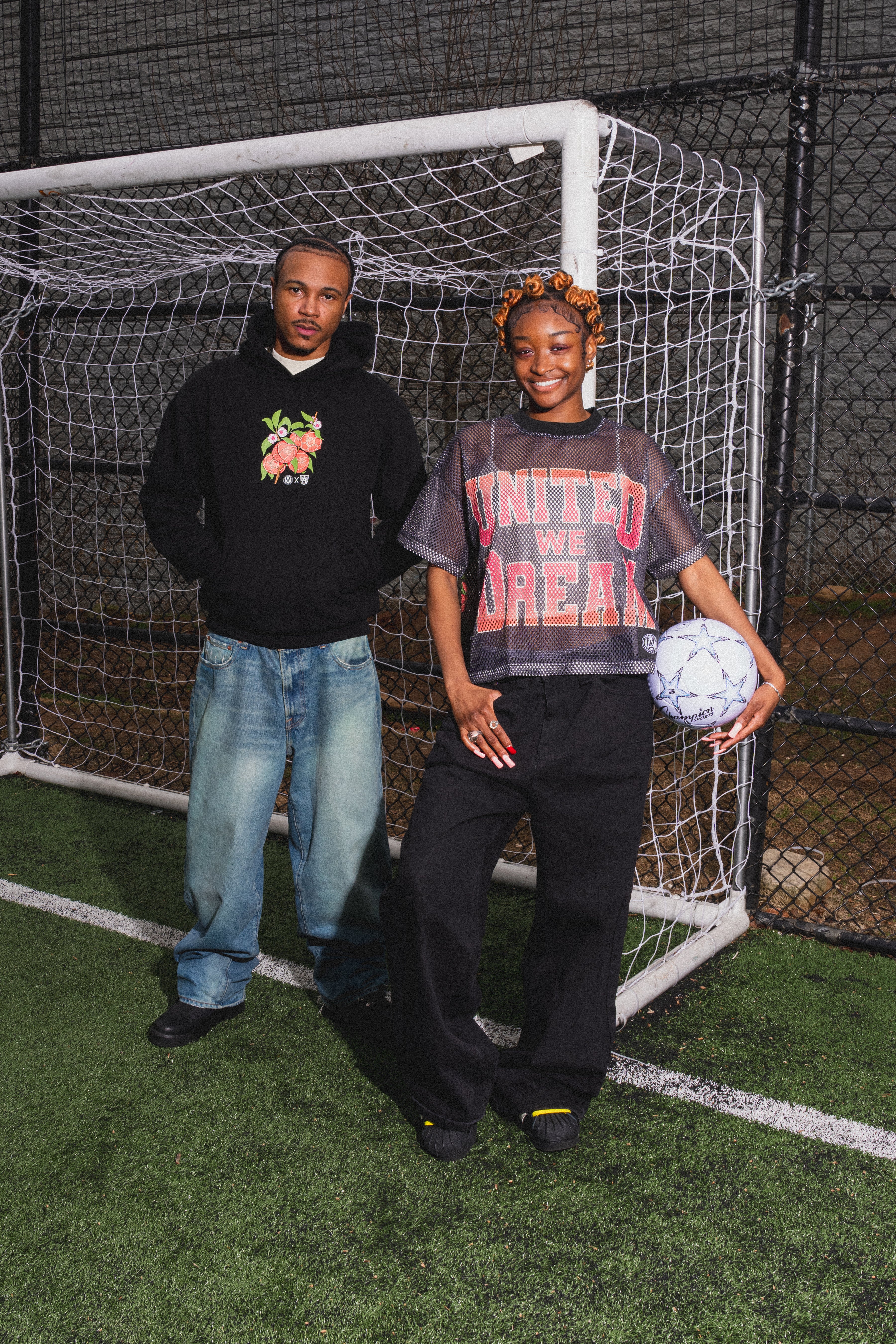 Two people standing in front of a soccer goal with a soccer ball.