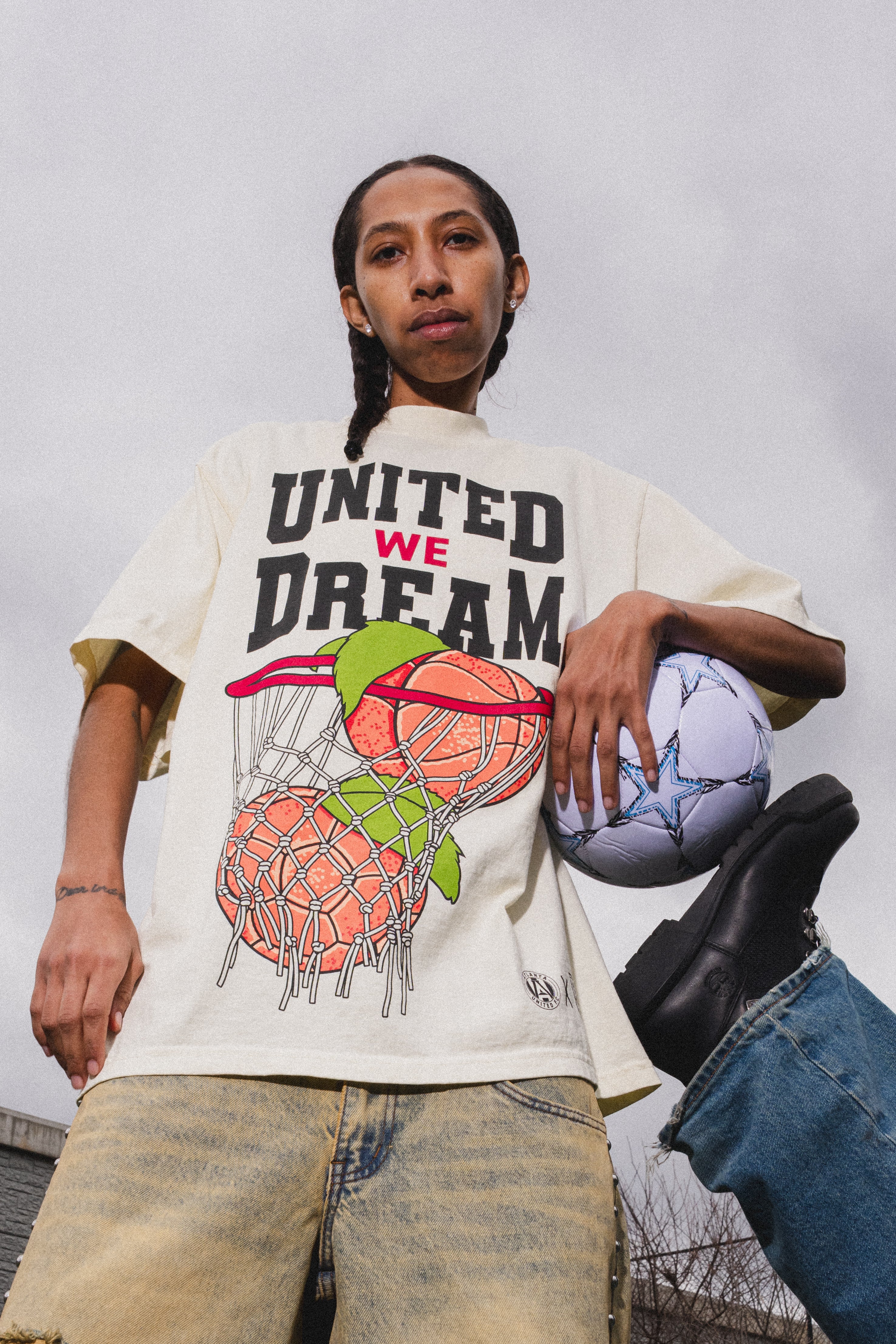 Person wearing a 'United We Dream' t-shirt with graphic design, holding a basketball and soccer ball.