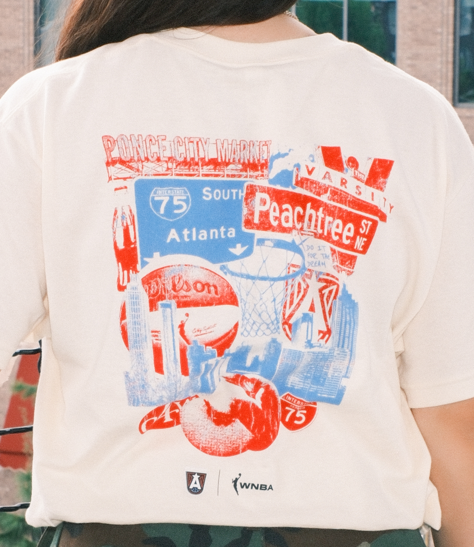 Close-up of the back of a beige t-shirt featuring a graphic with Atlanta landmarks, including Ponce City Market, Peachtree Street signs, a basketball hoop, a Wilson basketball, and the iconic peach. The graphic is in red and blue tones, with "Do It For The Dream" text and the Atlanta Dream logos at the bottom. The shirt is worn by a woman standing outdoors with camouflage pants.