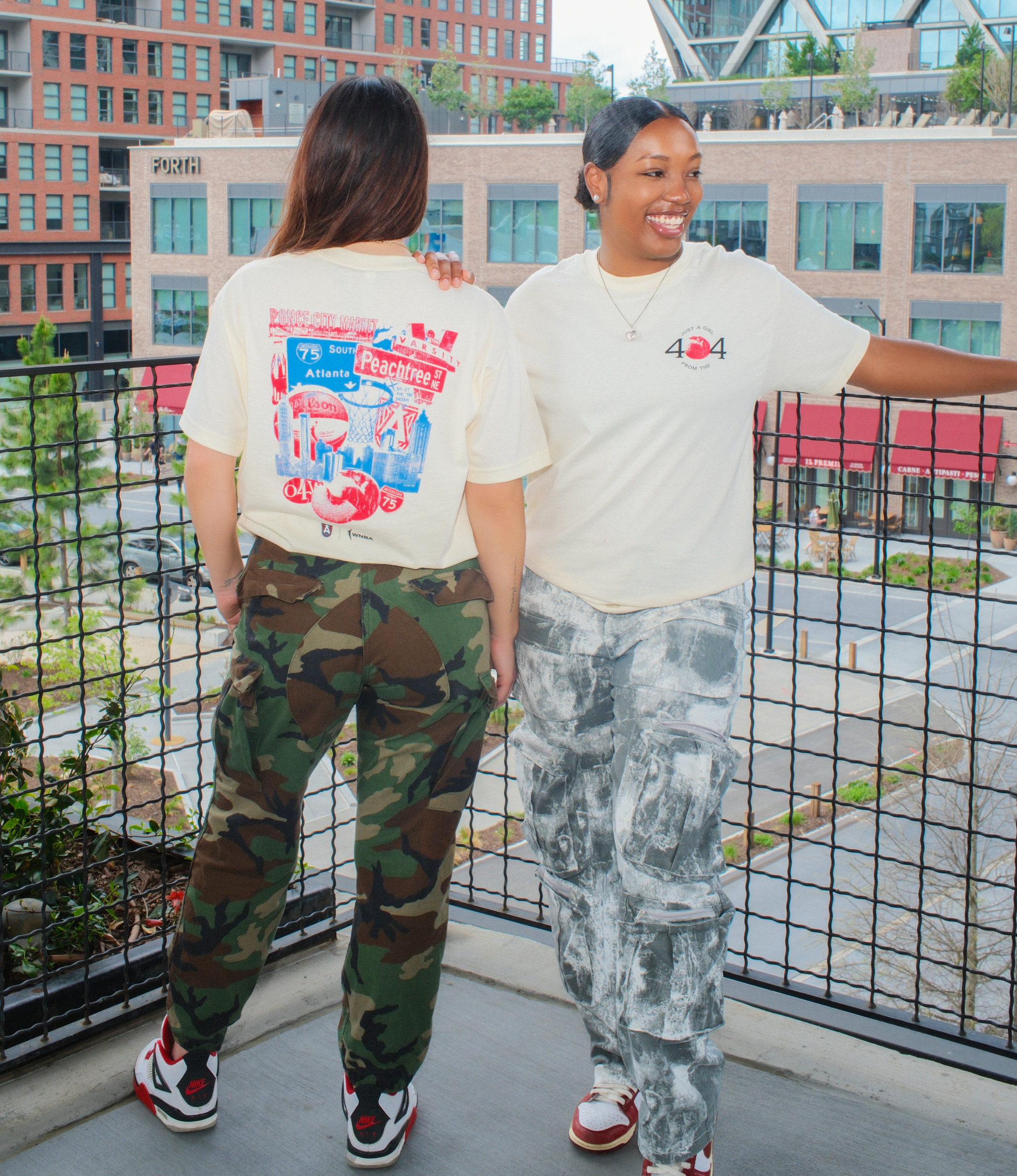 Two women standing side by side, both wearing the "Just A Girl From '404'" t-shirt. One woman is facing away, showing the back of the t-shirt with a graphic of Atlanta landmarks like Peachtree Street and a peach basketball. The other woman is smiling, wearing the front of the shirt with the "404" graphic. Both are dressed in casual pants—one in camouflage and the other in light gray cargo pants—paired with sneakers, against an urban background.