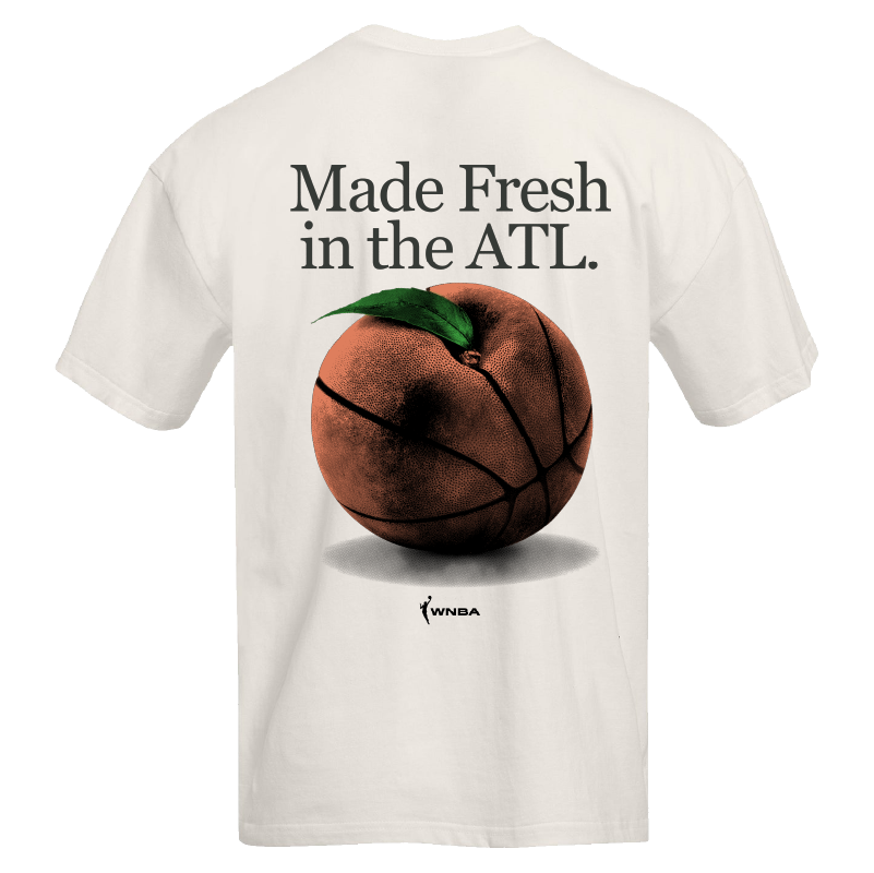 Atlanta Dream Made Fresh in ATL T-Shirt. White t-shirt featuring the graphic "Made Fresh in the ATL" in bold text above a basketball designed to look like an Atlanta peach. The shirt has a relaxed fit with drop shoulders, a raised collar, and a 1" neck band. The WNBA and Atlanta Dream logos are displayed at the bottom.